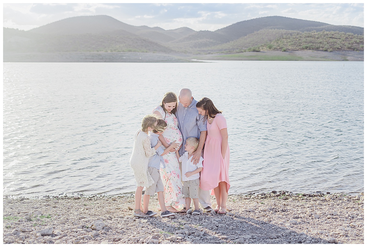 Family of six looks down at mom's pregnant belly during family maternity session at the lake