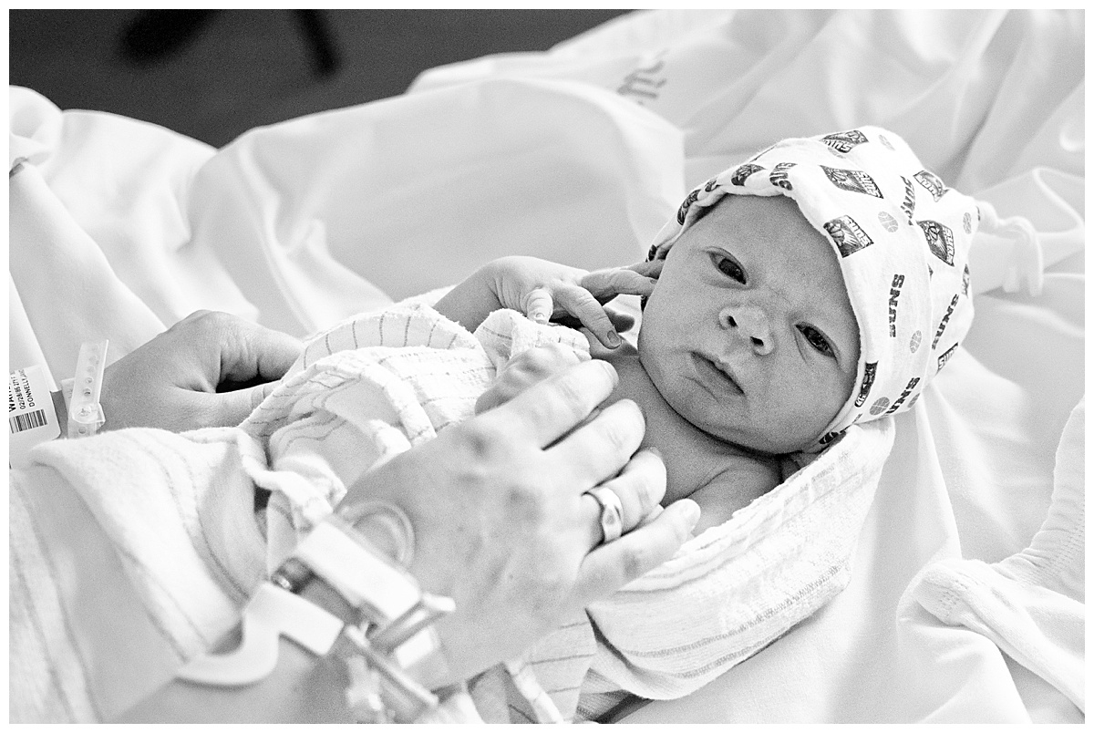 black and white image of newborn baby boy, swaddled and wearing a newborn cap in the hands of his mother
