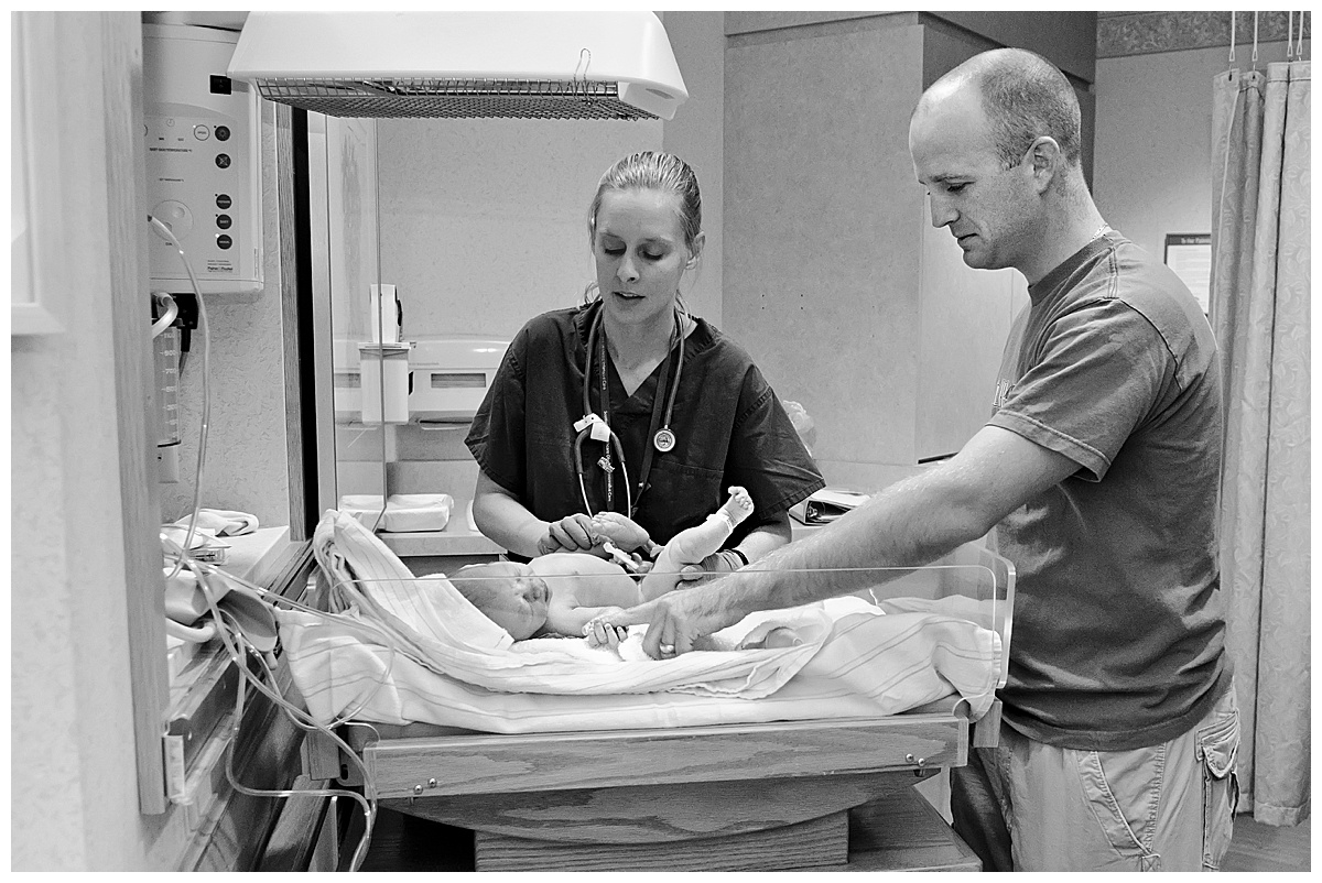 black and white image of a father looking on as his new baby is weighed by delivery room nurse