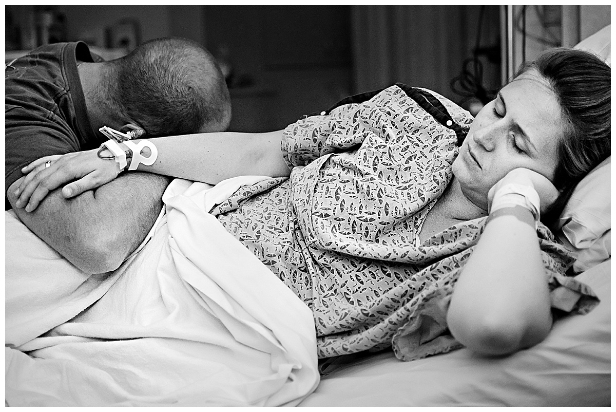 black and white image of mom in labor laying in hospital bed with husband resting on her side between contractions