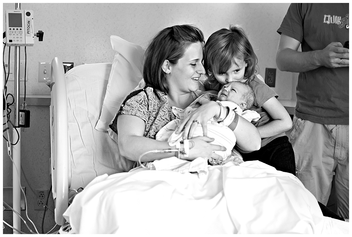 black and white image of big sister and mom holding and kissing brand new baby boy in Scottsdale Shea delivery room