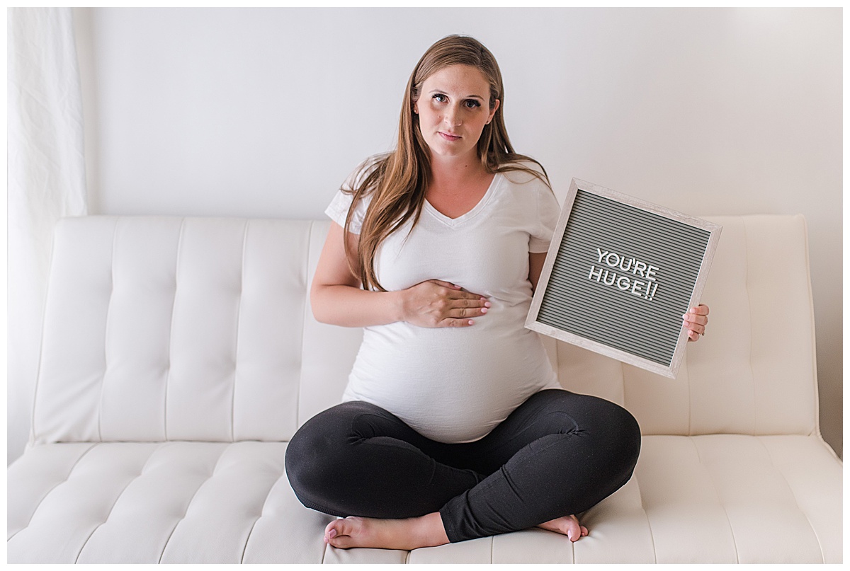 overdue pregnant woman sitting cross leg on a futon holding a letter board that says "You're huge!"