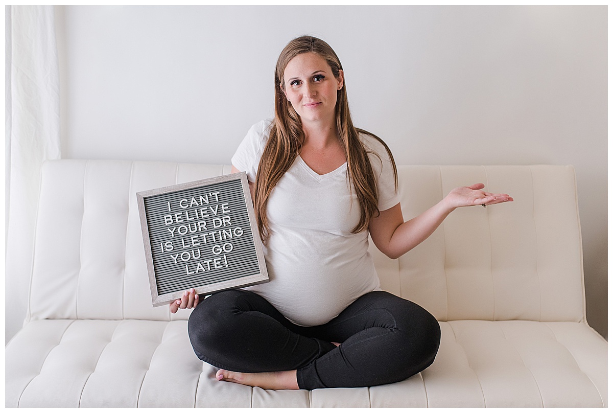 overdue pregnant woman sitting cross leg on a futon holding a letter board that says "I can't believe your dr is letting you go late"