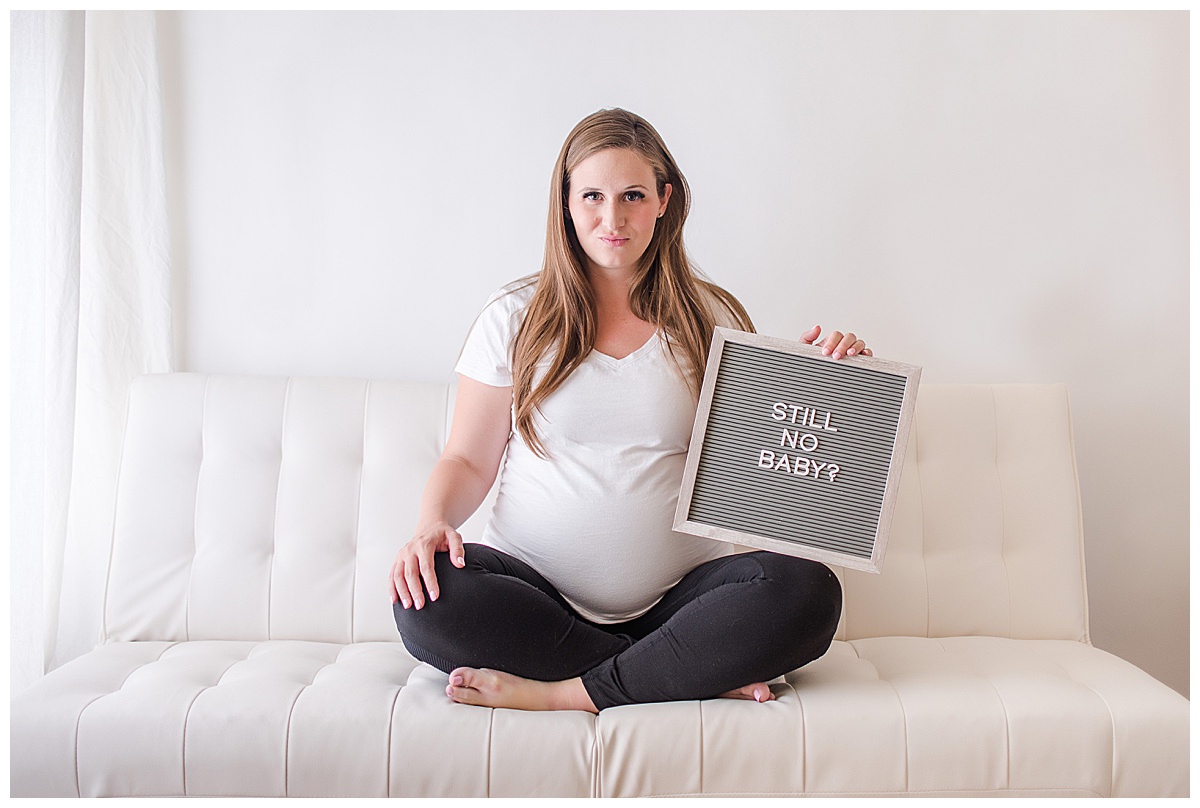 overdue pregnant woman sitting cross leg on a futon holding a letter board that says "Still no baby?"